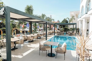 Un área de piscina exterior con sillas de descanso y mesas, rodeada por una zona cubierta con asientos y una piscina.