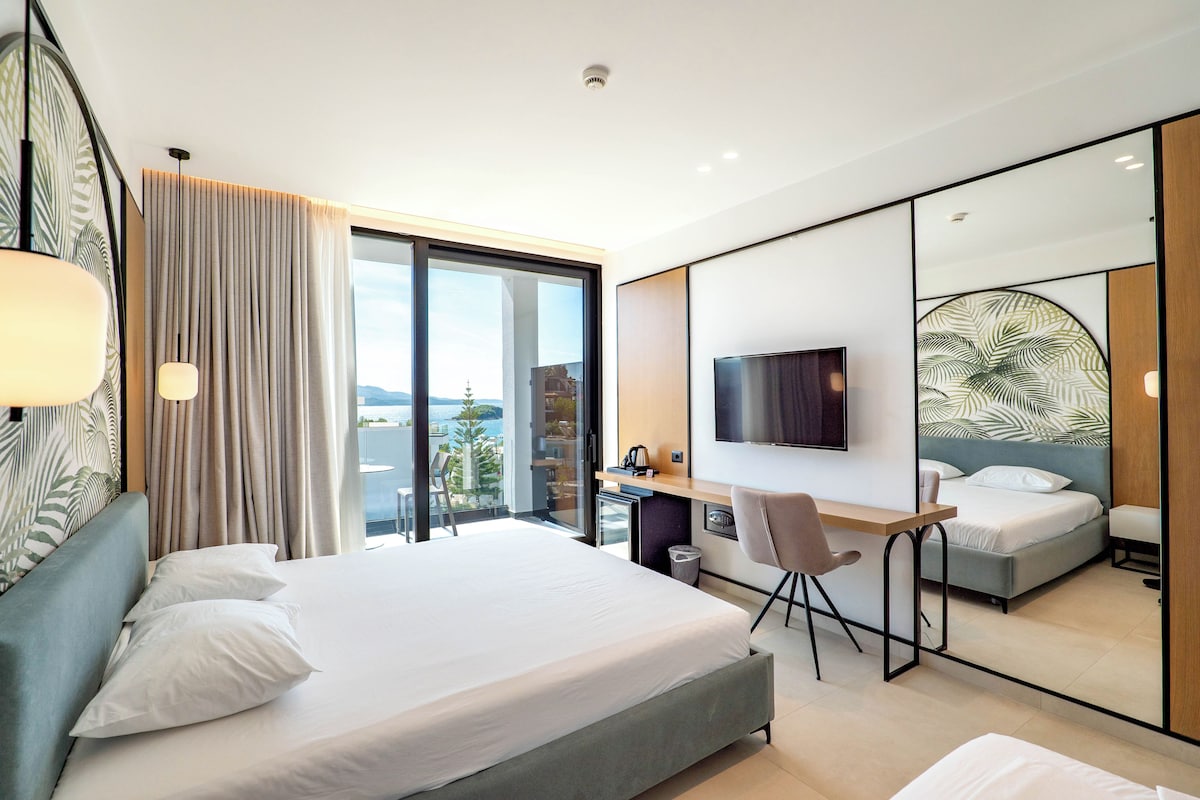 A modern hotel room with a large bed, a desk, a television, and a view of the sea.