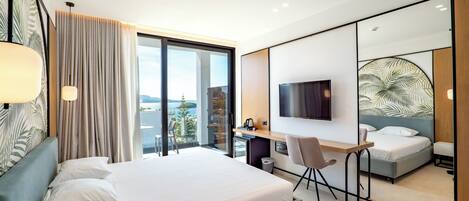 A modern hotel room with a large bed, a desk, a television, and a view of the sea.