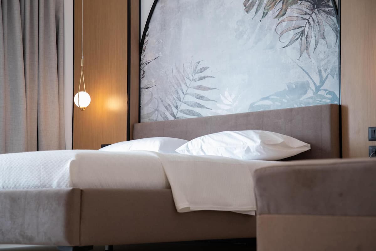 A neatly made bed with white linens, a hanging light fixture, and a large wall mural with leaf patterns.