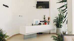 A reception area with a white reception desk, a flat-screen TV mounted on the wall, and a potted plant to the right.