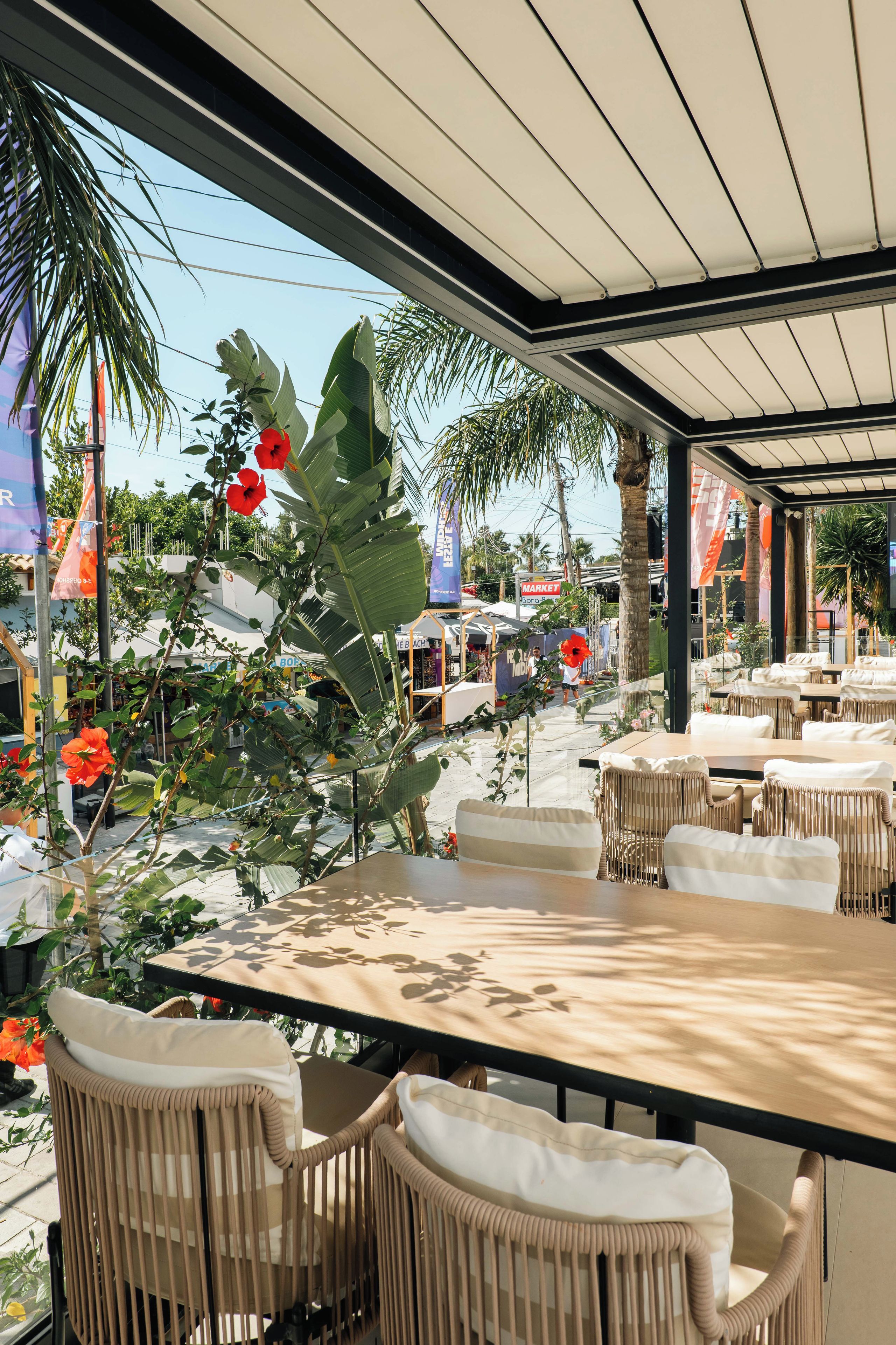 An outdoor cafe with tables and chairs, surrounded by tropical plants and a view of the street.