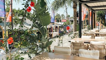 An outdoor cafe with tables and chairs, surrounded by tropical plants and a view of the street.