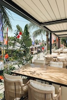 An outdoor cafe with tables and chairs, surrounded by tropical plants and a view of the street.