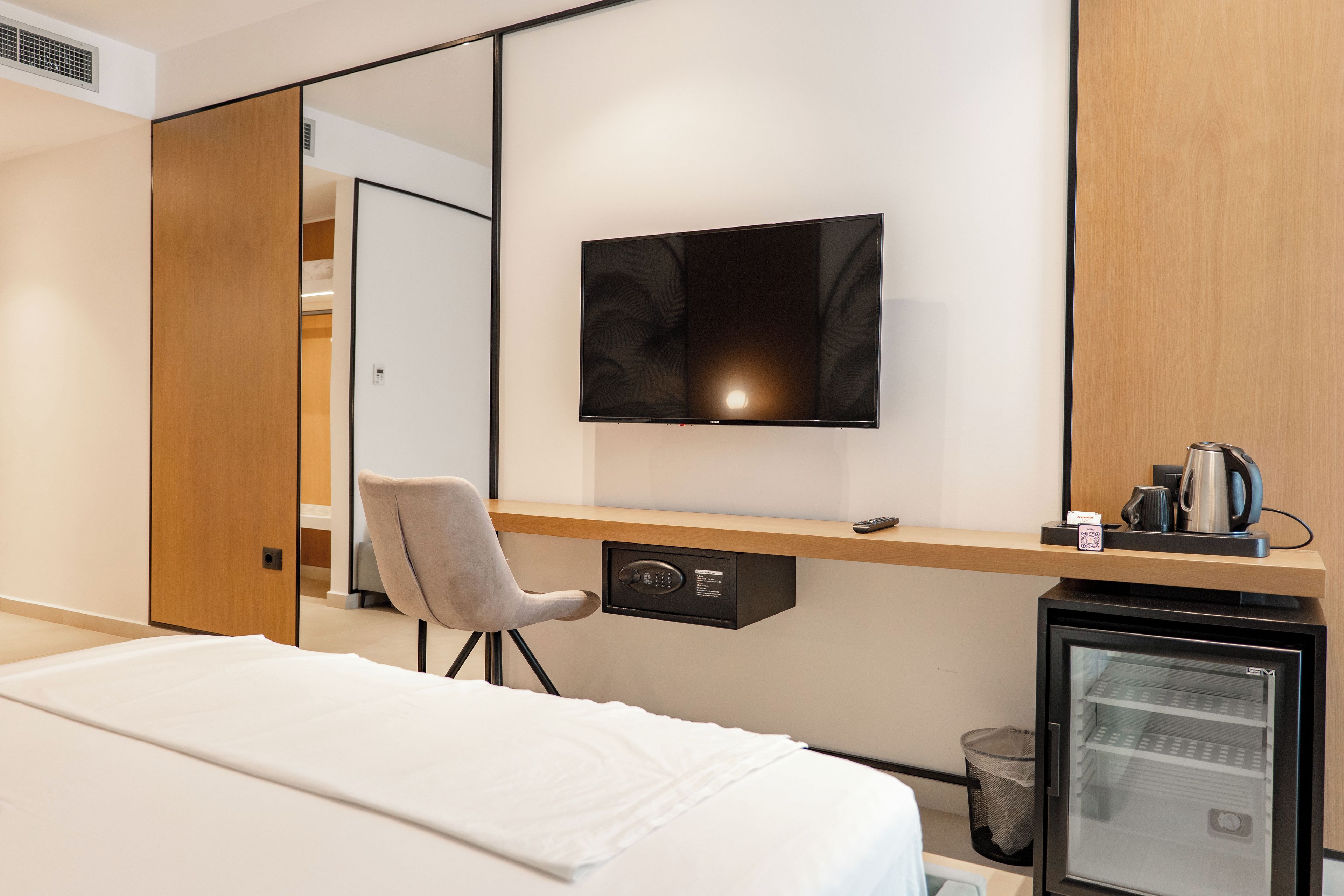 Una camera d'hotel con un letto, una scrivania con una sedia, una televisione a parete, una macchina per il caffè e un piccolo frigorifero.