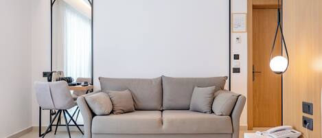 A modern hotel room with a neutral color scheme, a sofa, a desk, and a large mirror.
