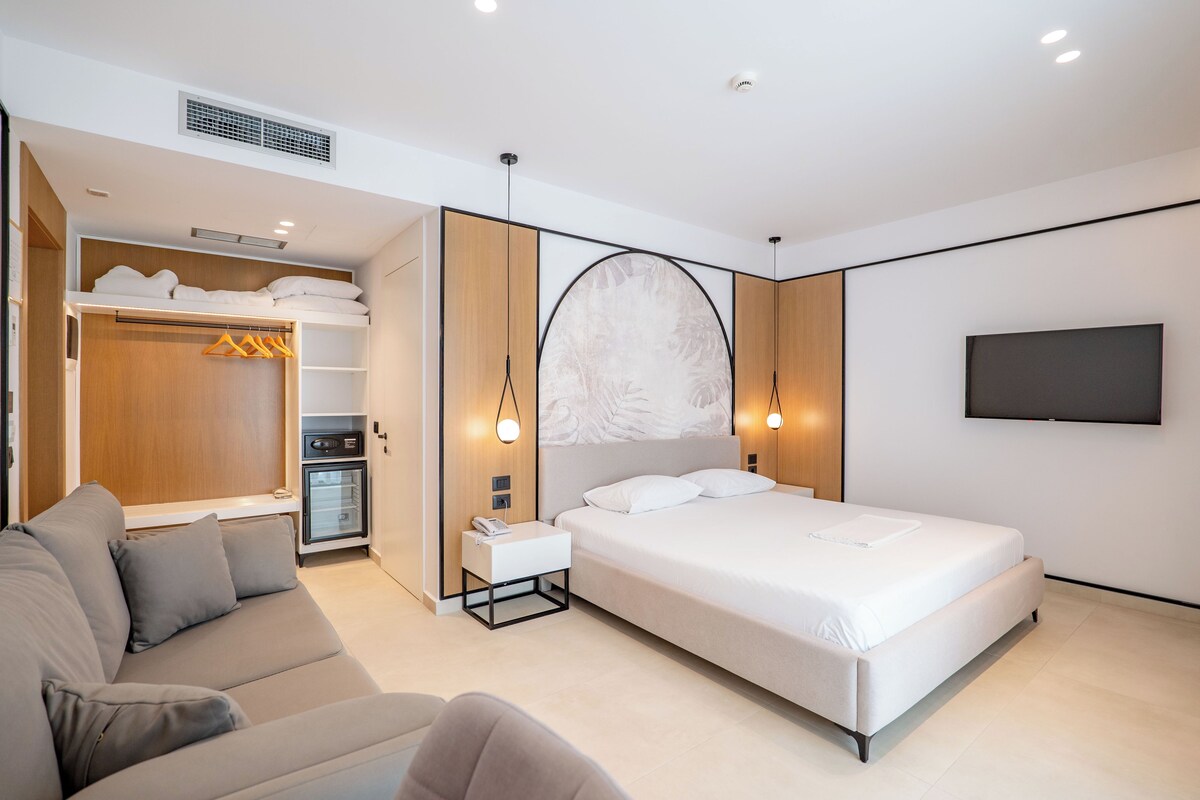 A modern hotel room with a bed, a sofa, a wardrobe, a minibar, and a TV mounted on the wall.