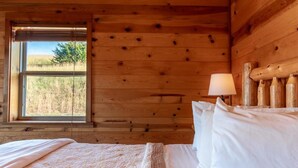 A wooden cabin room with a bed, a window with a view of the outdoors, and a lamp on the nightstand. - Mountain Sunset Cabin w/ Hot Tub (Ponca)