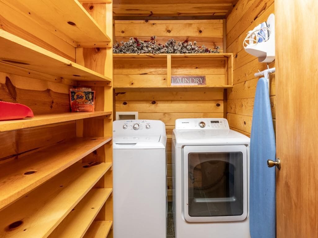Uma lavanderia com duas máquinas de lavar brancas, uma prateleira com detergente Tide e uma placa com a inscrição 'LAUNDRY'.