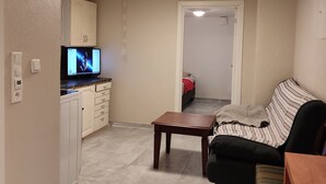 Sala de estar con sofá, mesa de centro y televisor al fondo. Se observa un área de cocina con armarios y un refrigerador.