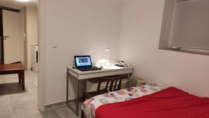 Un dormitorio con una cama, un escritorio con una computadora portátil, una silla y una ventana con persianas.