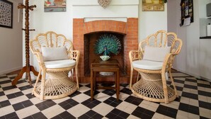 Een kamer met een zwart-witte vloer, twee rieten stoelen met kussens, een schoorsteen met pauwenmotieven, en een bordje met de tekst 'Welcome' erboven.