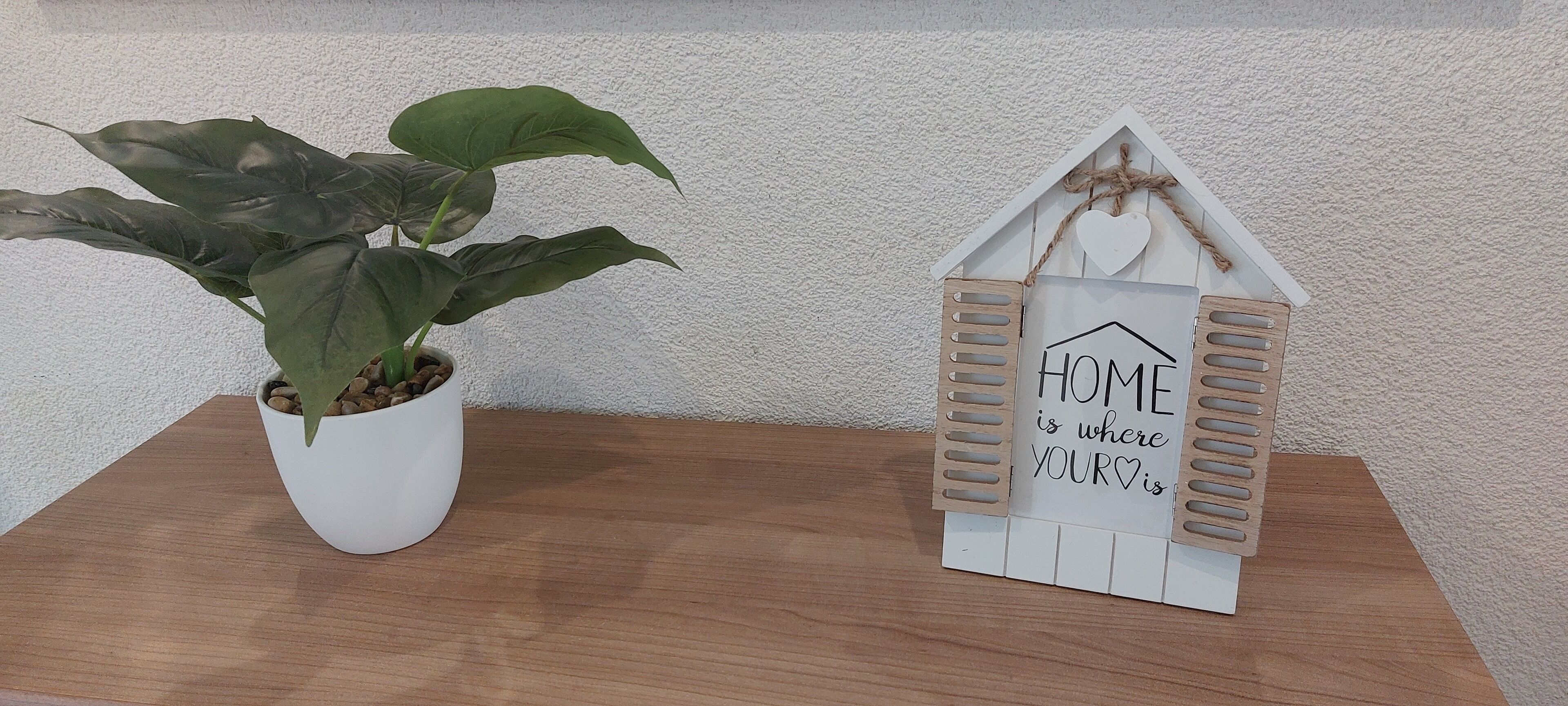 Ein Blumentopf und ein dekoratives Schild in Hausform mit dem Text 'HOME is where YOURS is' auf einer Holzoberfläche vor einer strukturierten Wand.