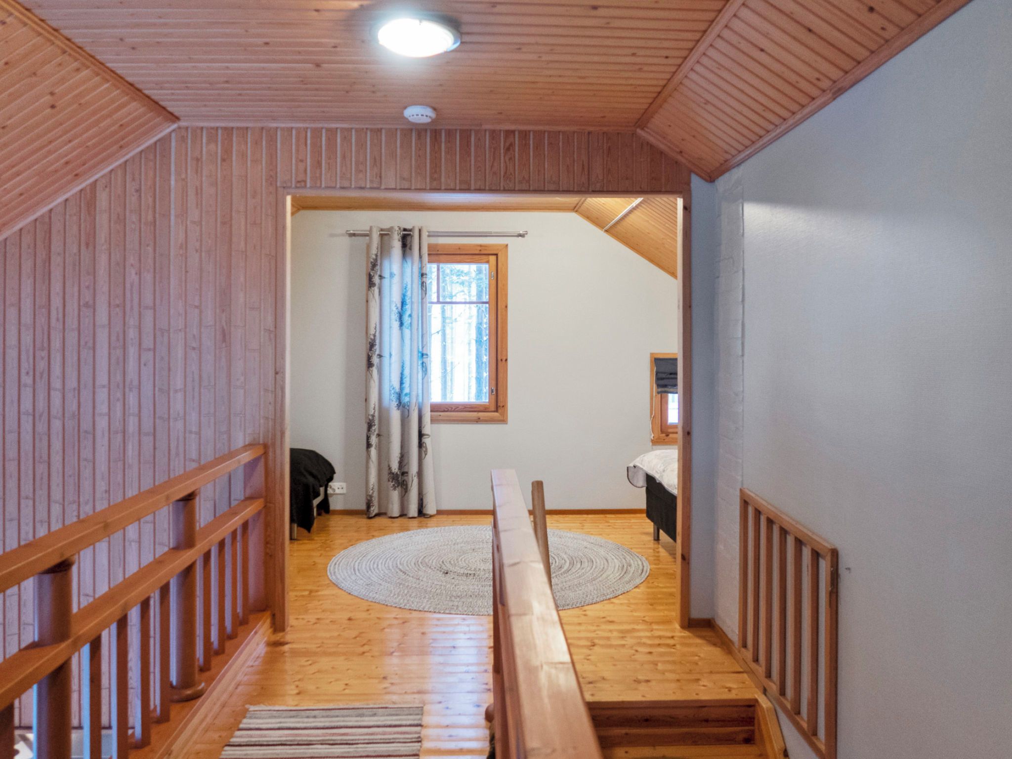 Ein Holzinnenraum mit einer Treppe, einem Fenster mit Vorhängen und einem Bett mit Nachttisch.
