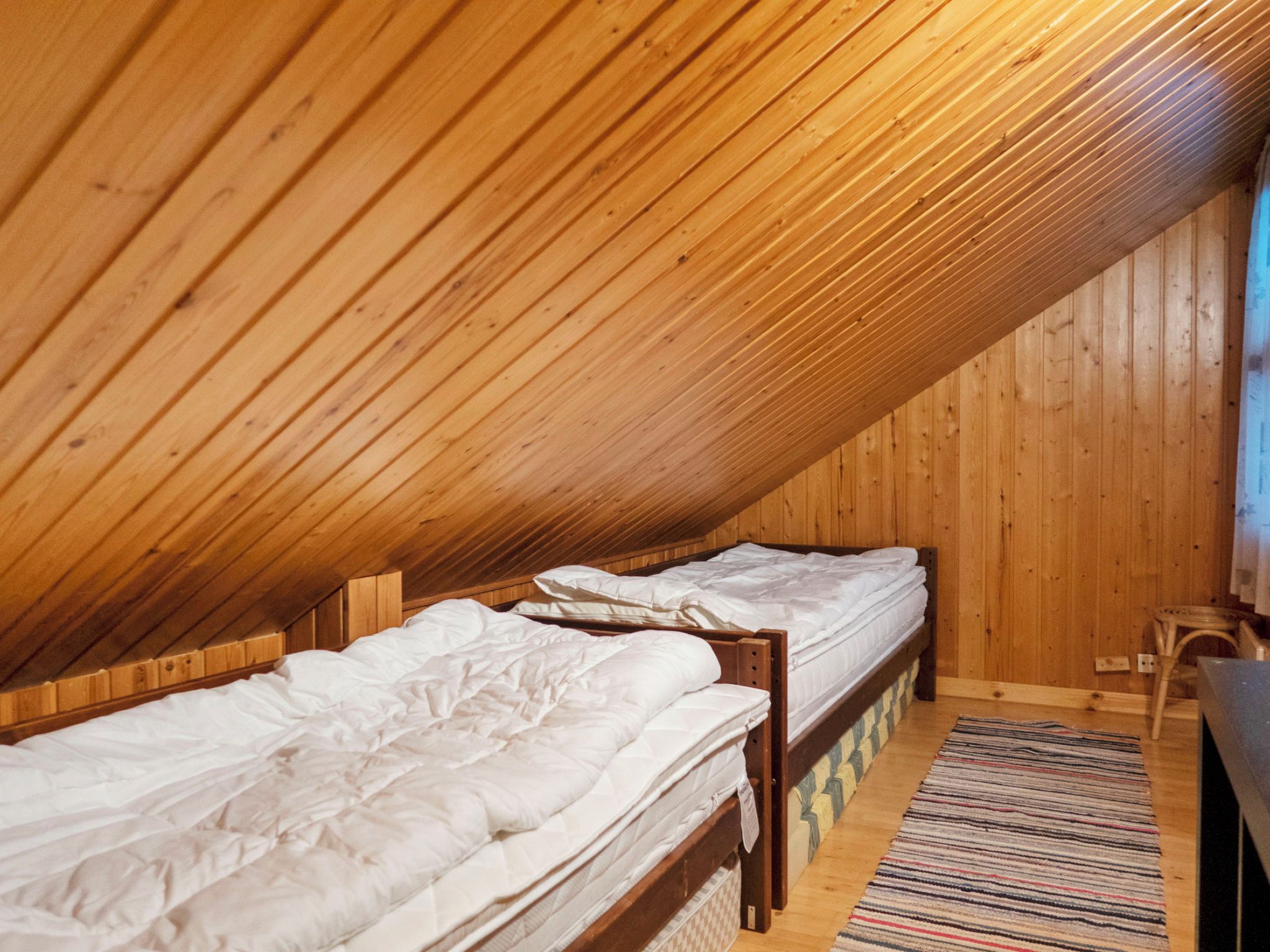A small, cozy attic bedroom with wooden slats on the ceiling, two single beds, a small table, and a chair.