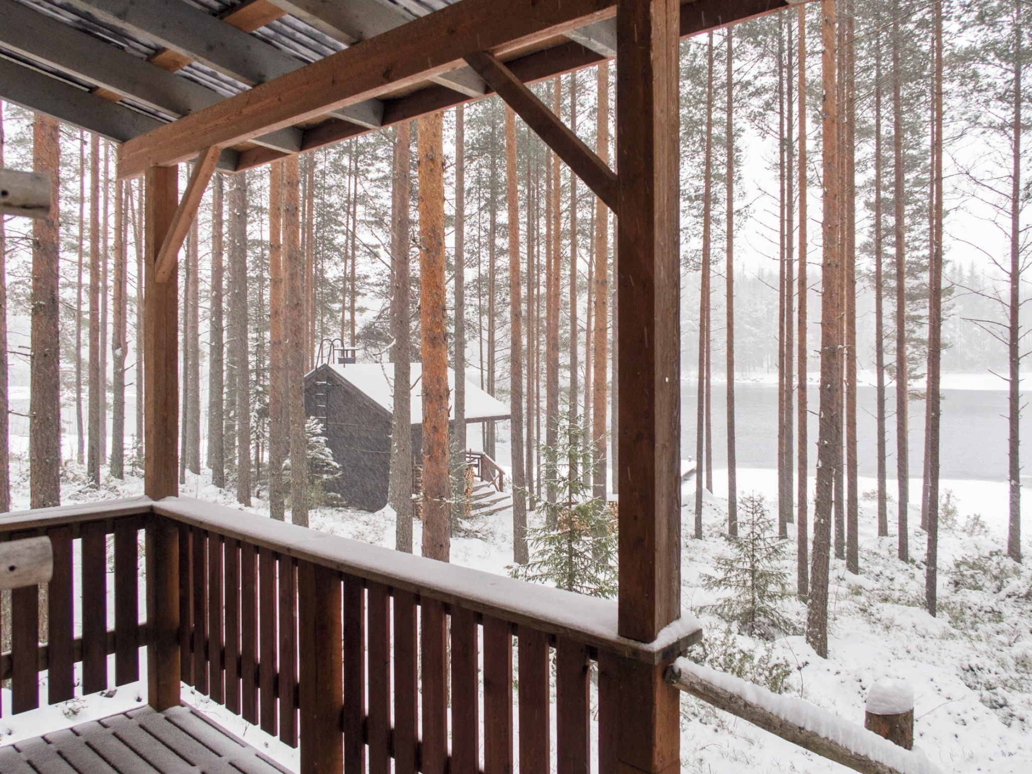 A wooden deck with a view of a cabin surrounded by tall trees and a snowy landscape.