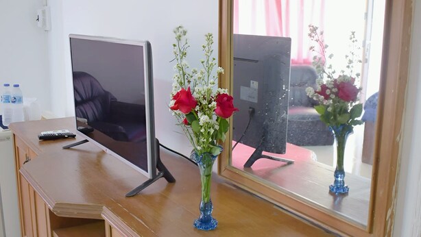 Un escritorio de madera con un televisor, un florero con flores y un espejo que refleja una habitación con una cortina rosa.