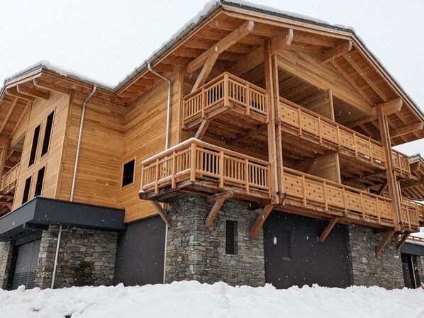 A wooden chalet with a stone foundation, multiple balconies, and a snow-covered roof.