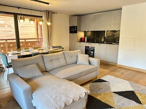 Una sala de estar moderna con un sofá y un comedor con mesa dispuesta para cuatro. Se ve una cocina al fondo con encimera negra y armarios blancos.