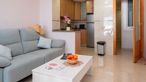 A living room with a grey sofa, a white coffee table with a bowl of fruit, and a magazine. A kitchen is visible in the background with wooden cabinets and a microwave. - Modern Apartment Facing Sant Antoni Beach (San Antonio de Calonge)