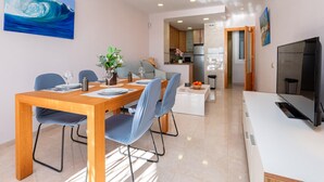A modern dining area with a wooden table and blue chairs, a kitchen in the background, and a TV on the right. - Modern Apartment Facing Sant Antoni Beach (San Antonio de Calonge)