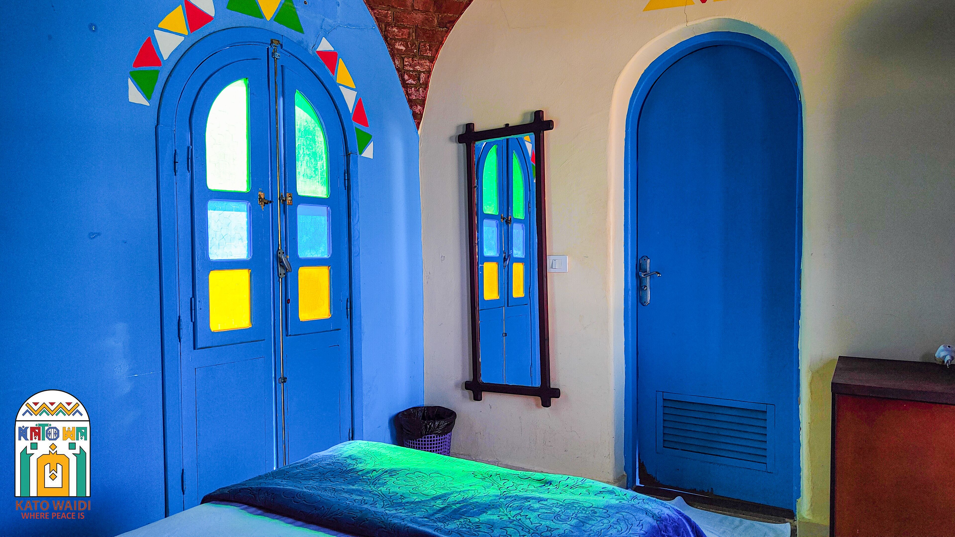 Een kamer met twee blauwe deuren, waarvan er eentje een spiegel heeft, een bed met een bedspread met een groen en blauw patroon, en een kleine prullenbak.