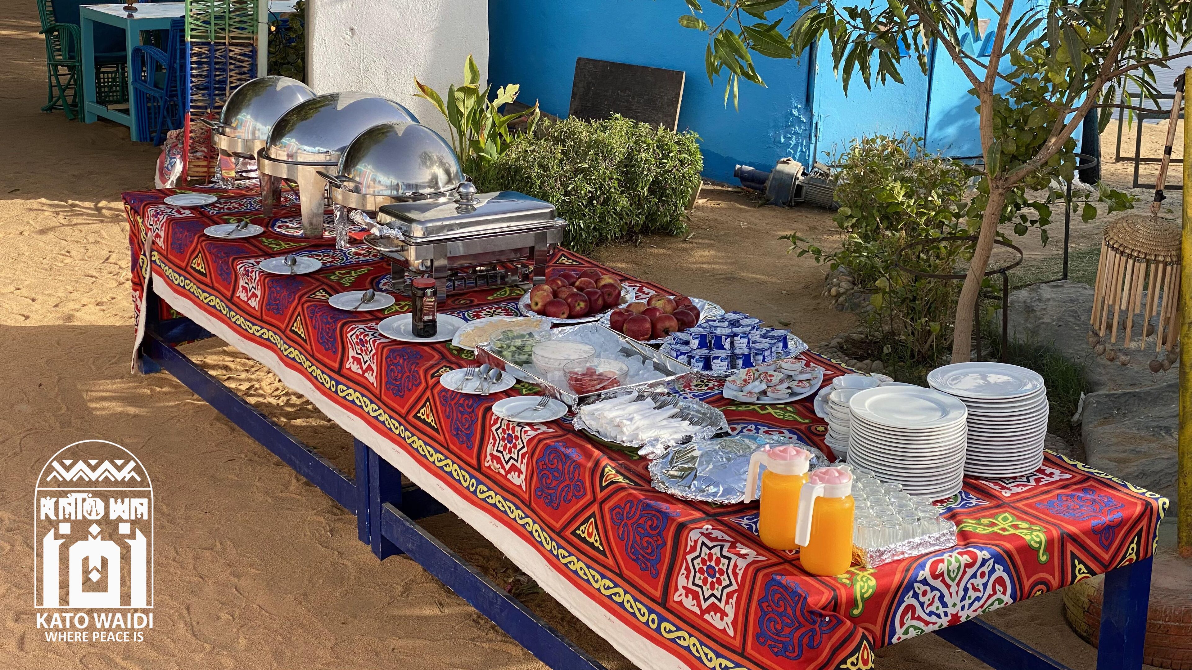 A buffet setup with various food items and utensils, a blue wall in the background, and a decorative tree.