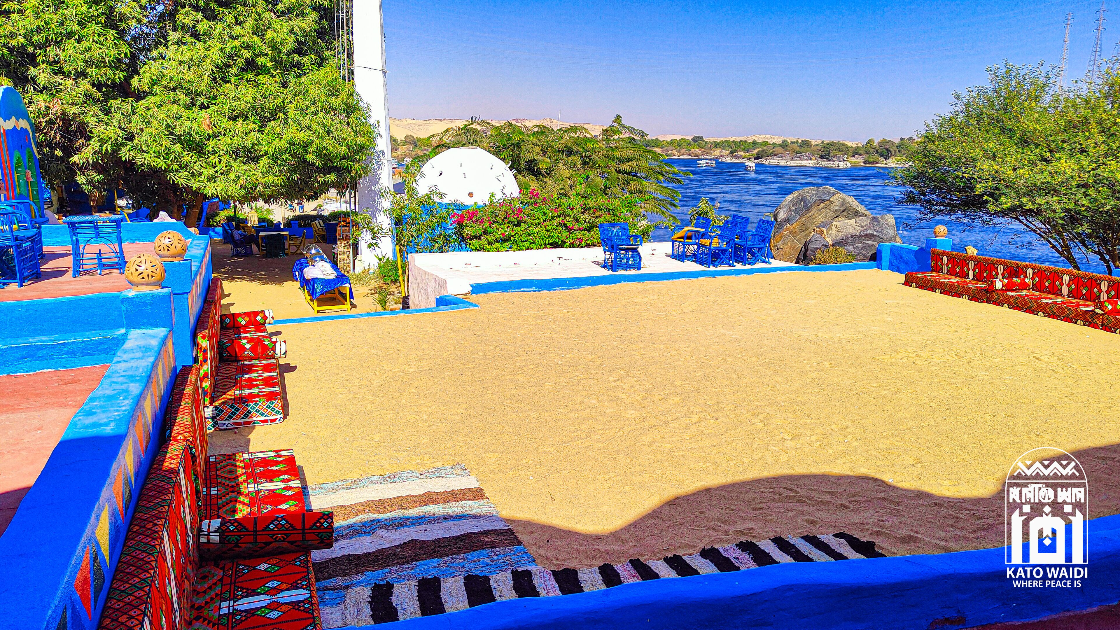 Ein Strand mit blauer und roter Sitzgelegenheit, einer weißen Kuppelkonstruktion und Blick auf das Meer und die Hügel.