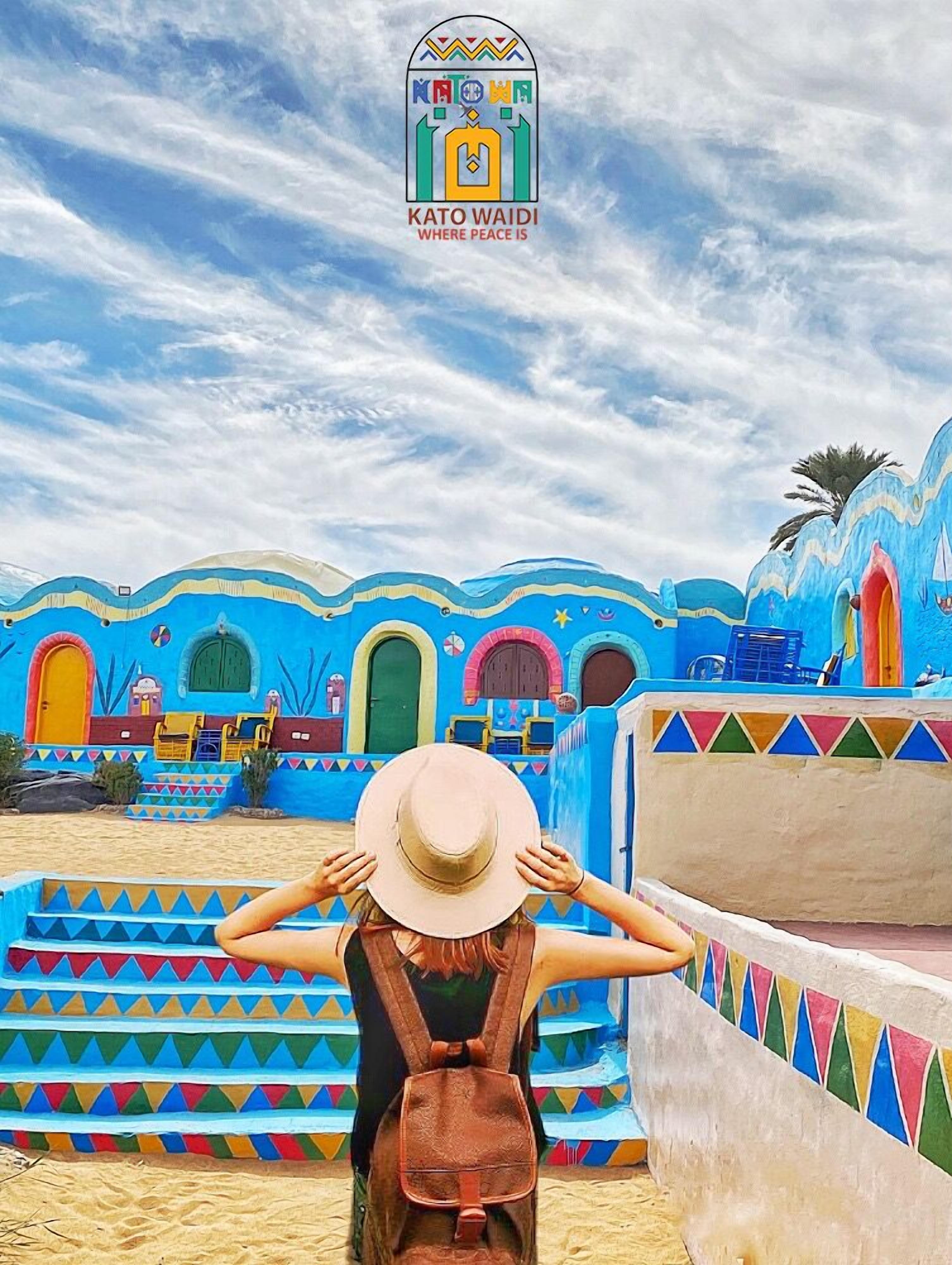 A person standing in front of a colorful building with a logo reading "KATO WAIDI WHERE PEACE IS".