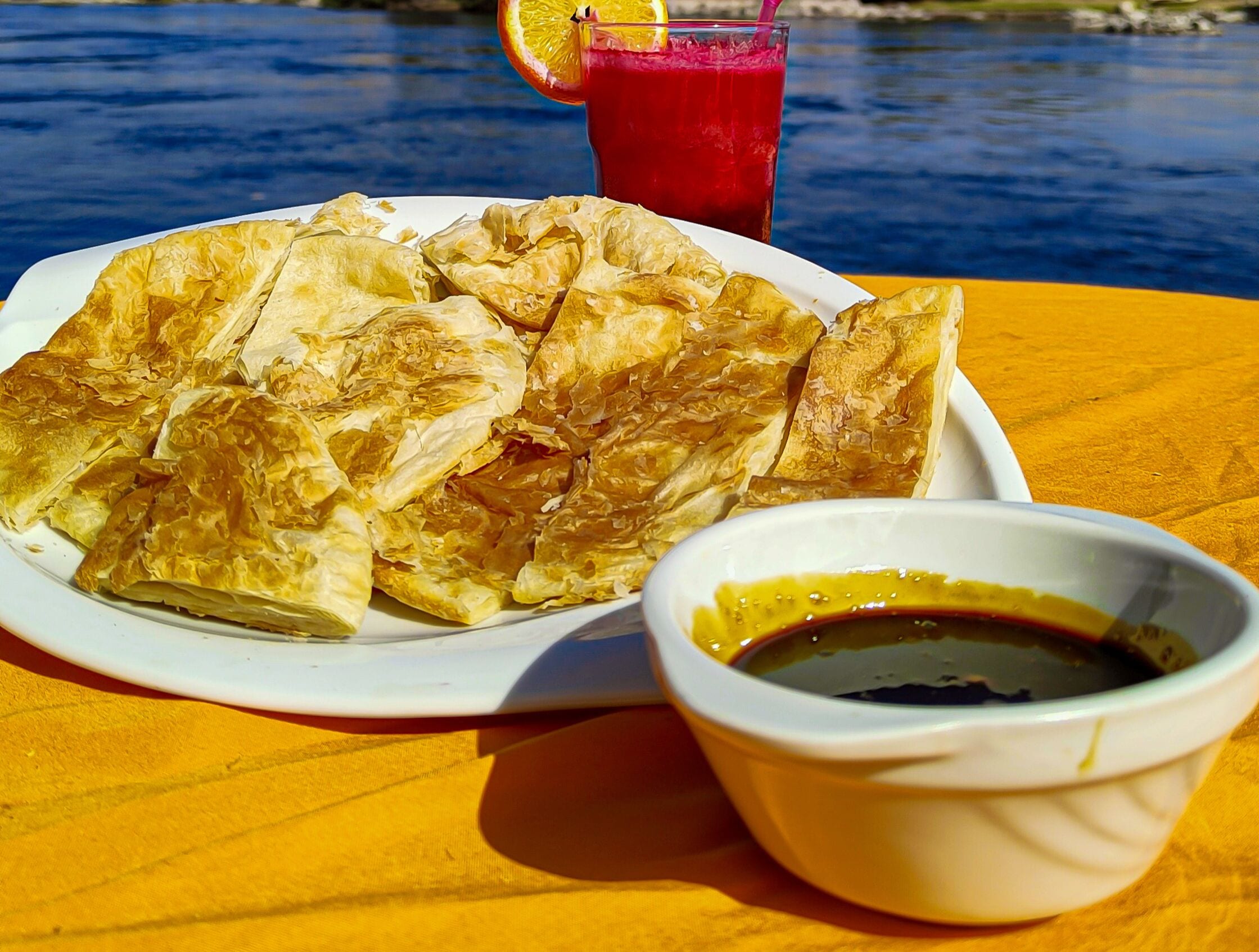 A plate of phyllo pastry with a dipping sauce, a glass of red drink, and a slice of orange on a yellow tablecloth by a body of water.