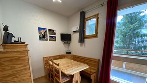 Una habitación con una mesa de madera y bancos, un televisor en la pared y una ventana con vista a árboles.