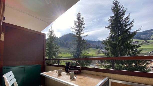 Un balcón con mesa y sillas, con vista a un paisaje montañoso con árboles y un pueblo.