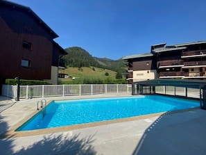 A swimming pool with a white fence, a building with balconies, and a mountainous landscape in the background.