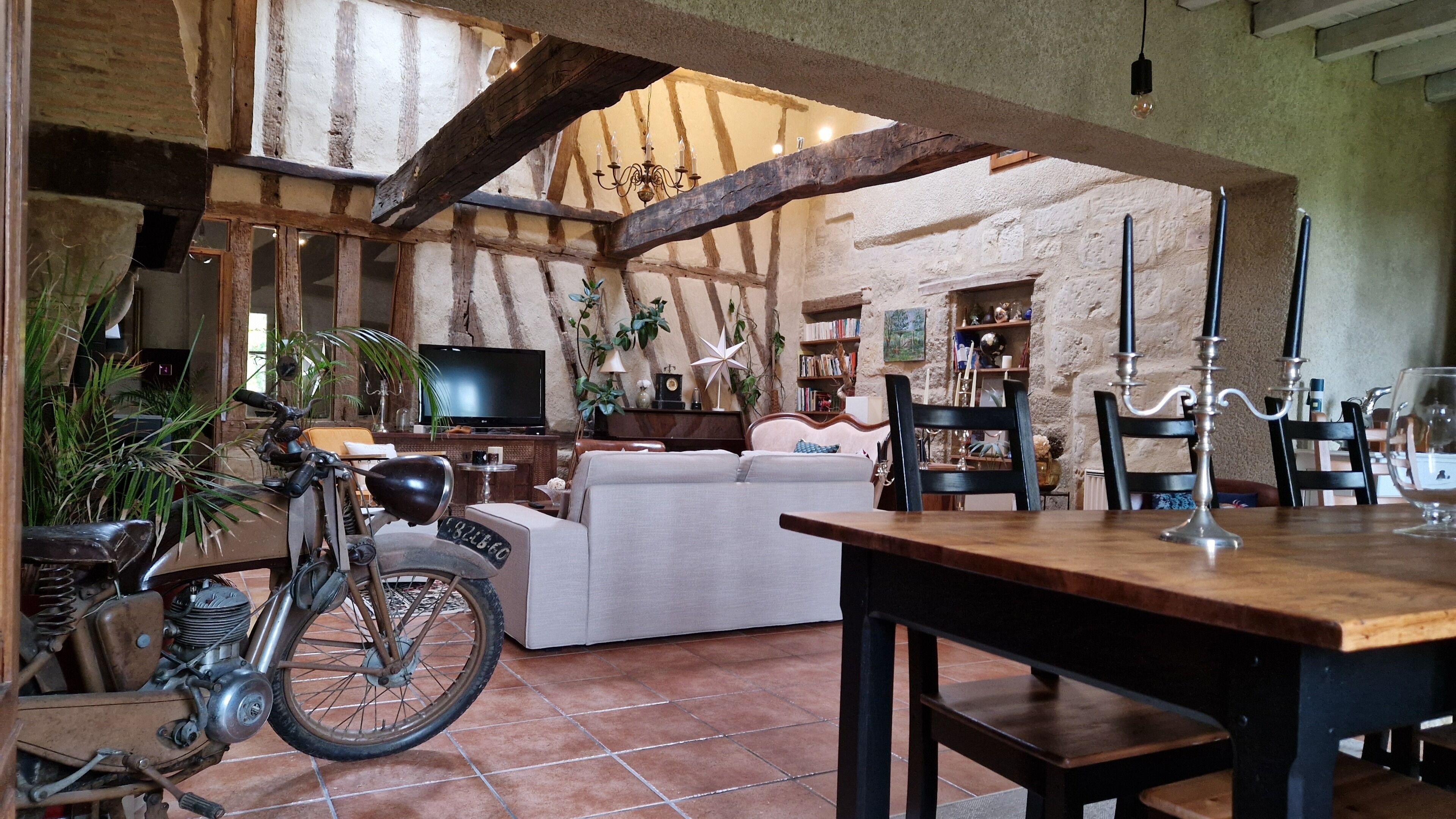A rustic interior with exposed wooden beams, a dining table, a sofa, and a vintage motorcycle.