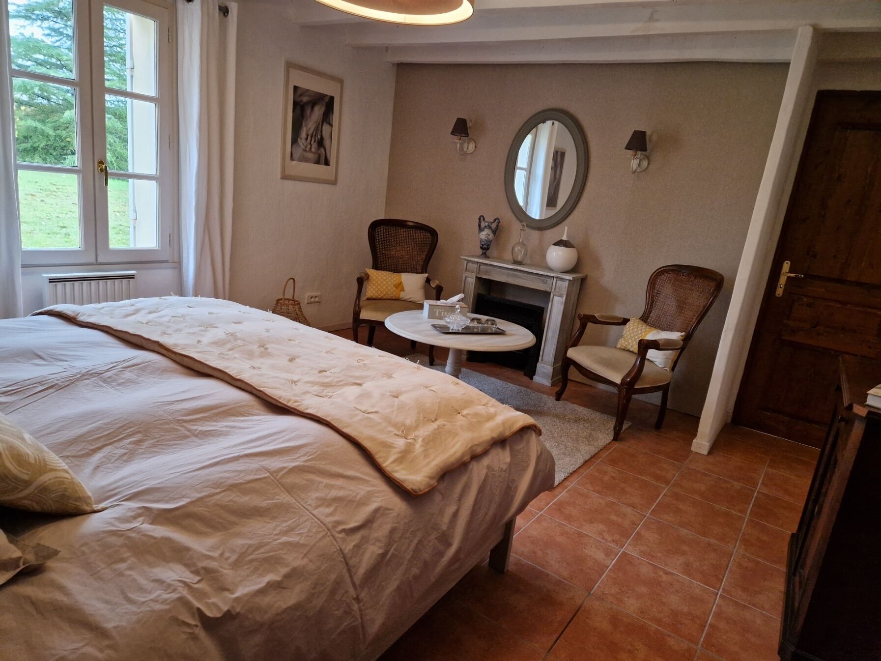 A bedroom with a large bed, two chairs, a small table, a fireplace, and a mirror on the wall.