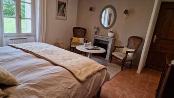 A bedroom with a large bed, two chairs, a small table, a fireplace, and a mirror on the wall.