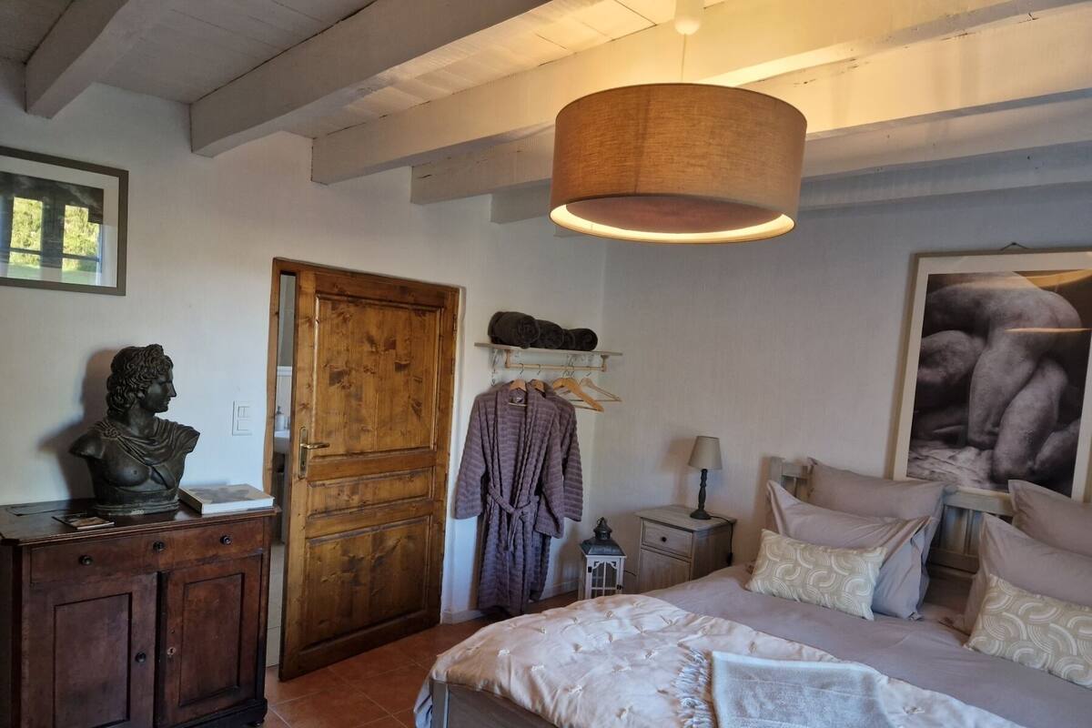A bedroom with a bed, a wooden wardrobe, a door, a nightstand, a lamp, and a framed picture on the wall.