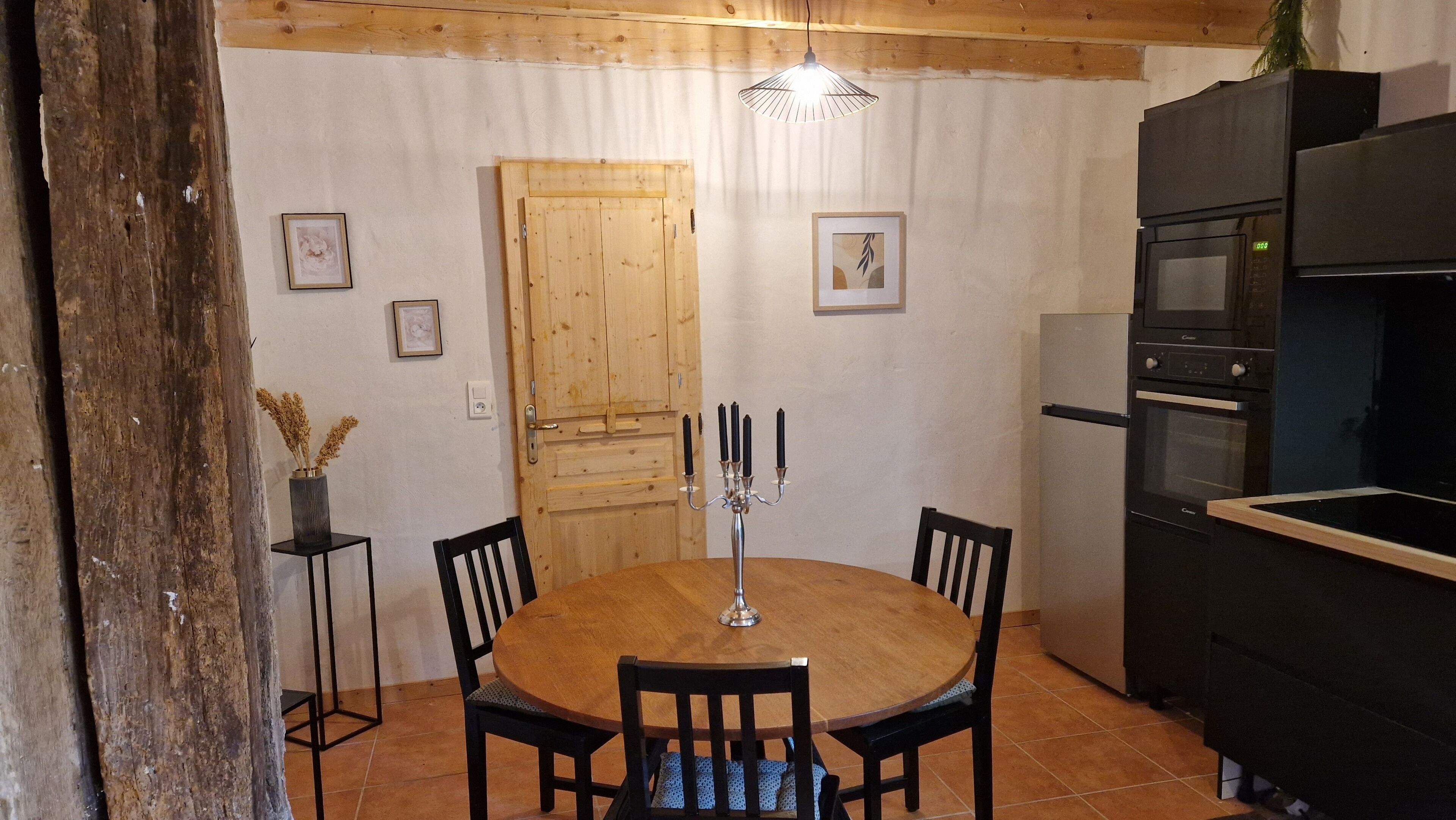 Une table à manger en bois, accompagnée de quatre chaises, d’un chandelier et d’un luminaire suspendu, dans un coin cuisine doté de meubles en bois et d’un réfrigérateur.
