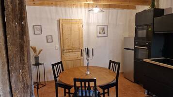 Une table à manger en bois, accompagnée de quatre chaises, d’un chandelier et d’un luminaire suspendu, dans un coin cuisine doté de meubles en bois et d’un réfrigérateur.