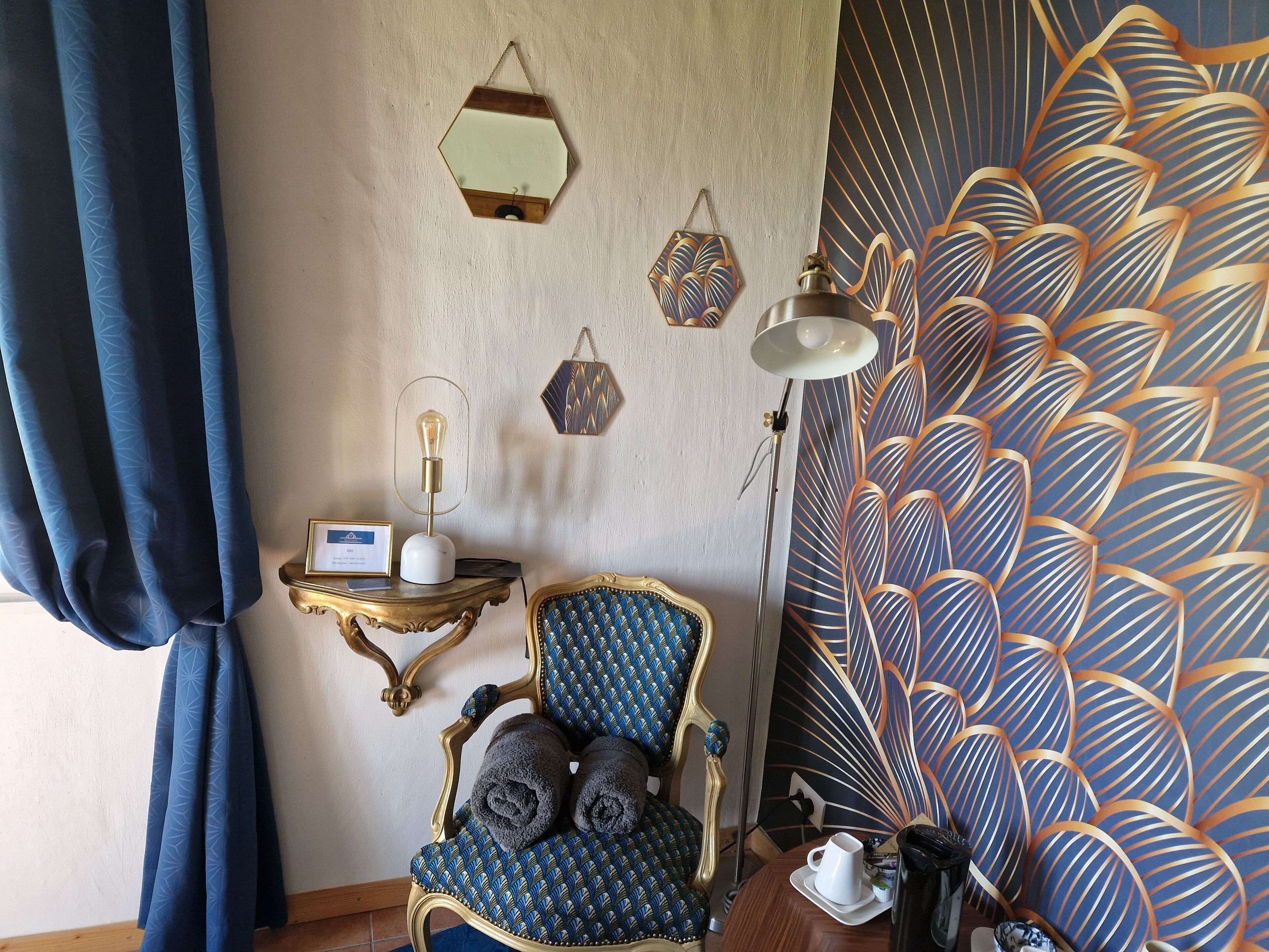 A room with a patterned wall, a chair with rolled towels, a table with a lamp and a sign, and a decorative mirror.