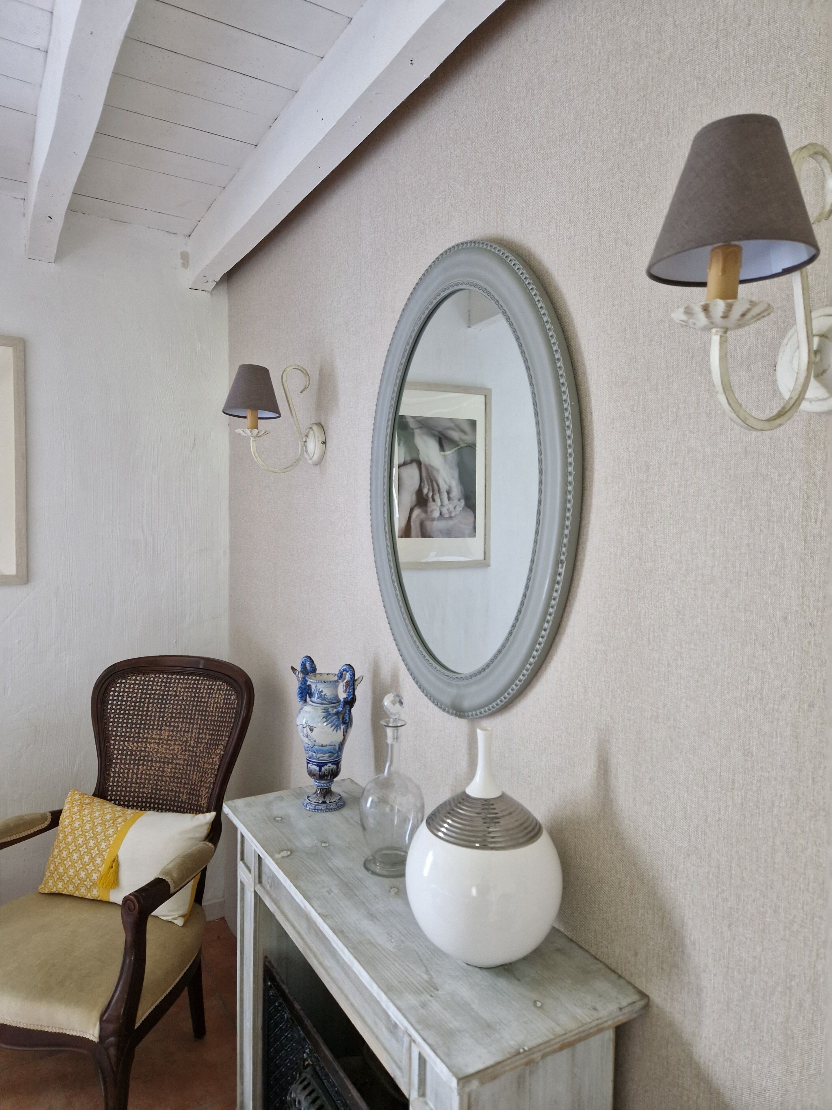 A room with a wooden chair, a marble fireplace, a wall-mounted mirror, and a wall sconce.