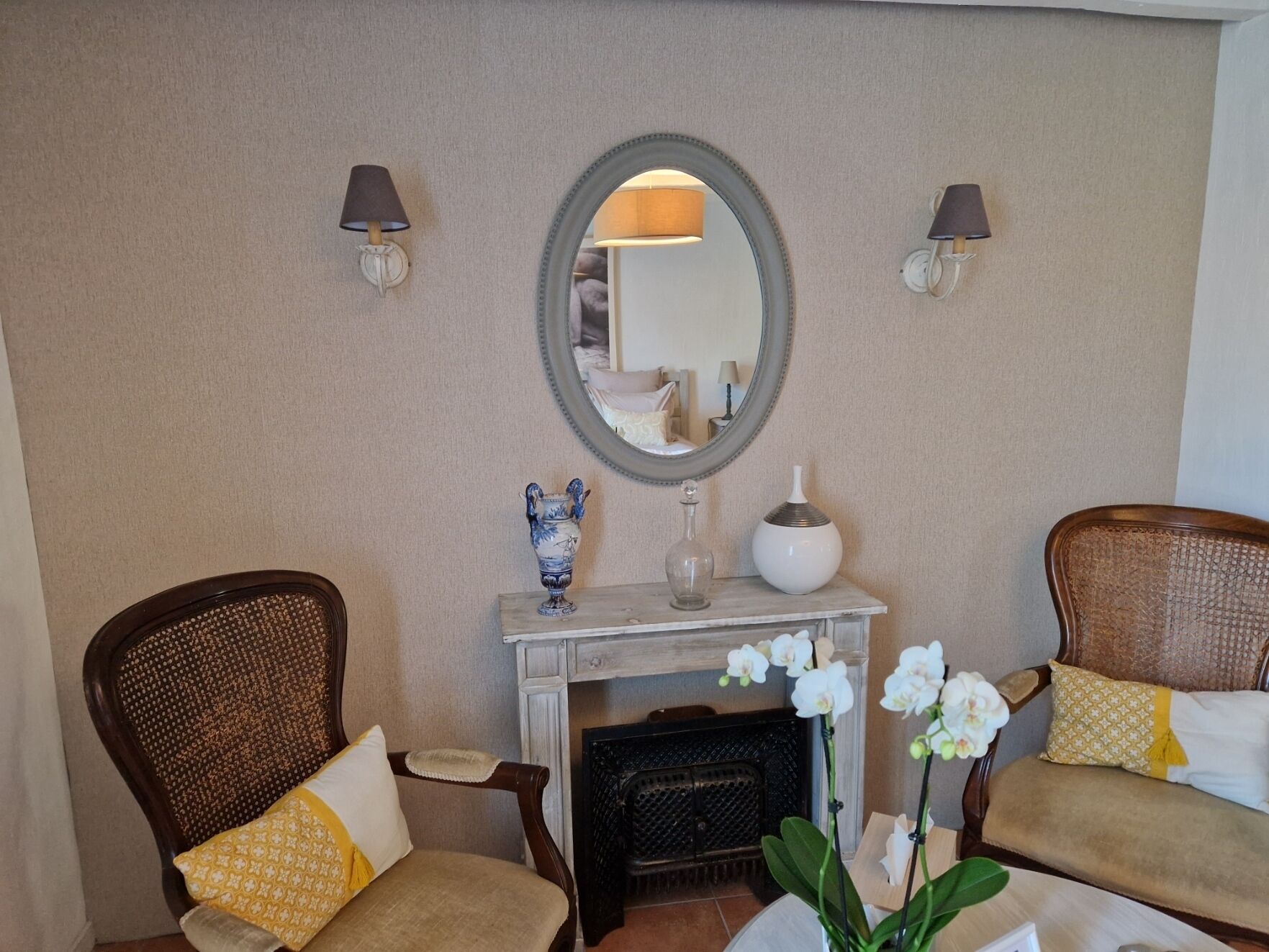 Une pièce comprenant deux fauteuils en osier, une console blanche agrémentée d’objets décoratifs, une cheminée et un grand miroir mural.