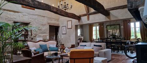 A spacious living room with a high ceiling, exposed wooden beams, a chandelier, and a dining area with a table and chairs.