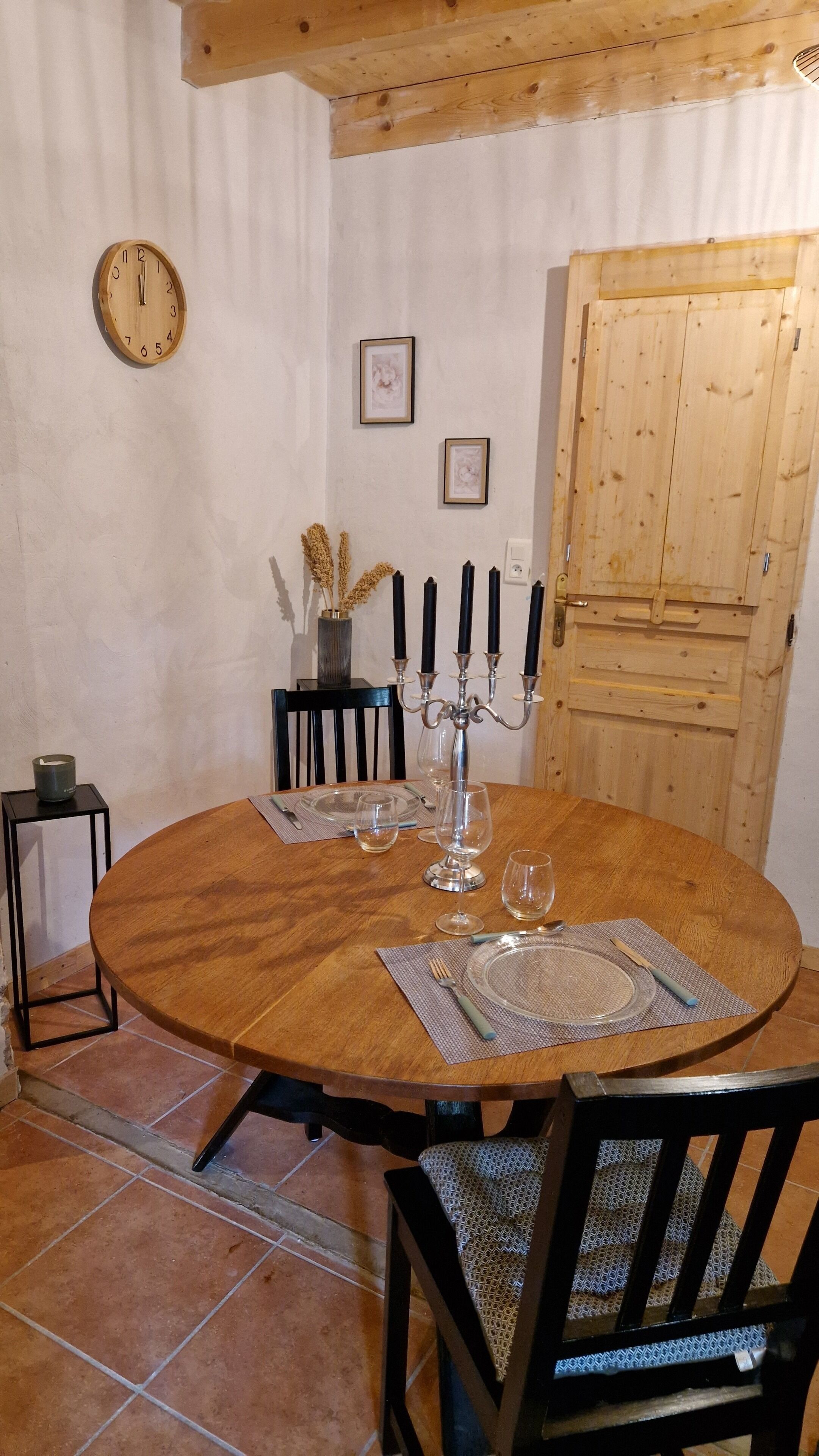 A wooden dining table with a candelabra, wine glasses, and cutlery. There is a clock on the wall and a door in the background.