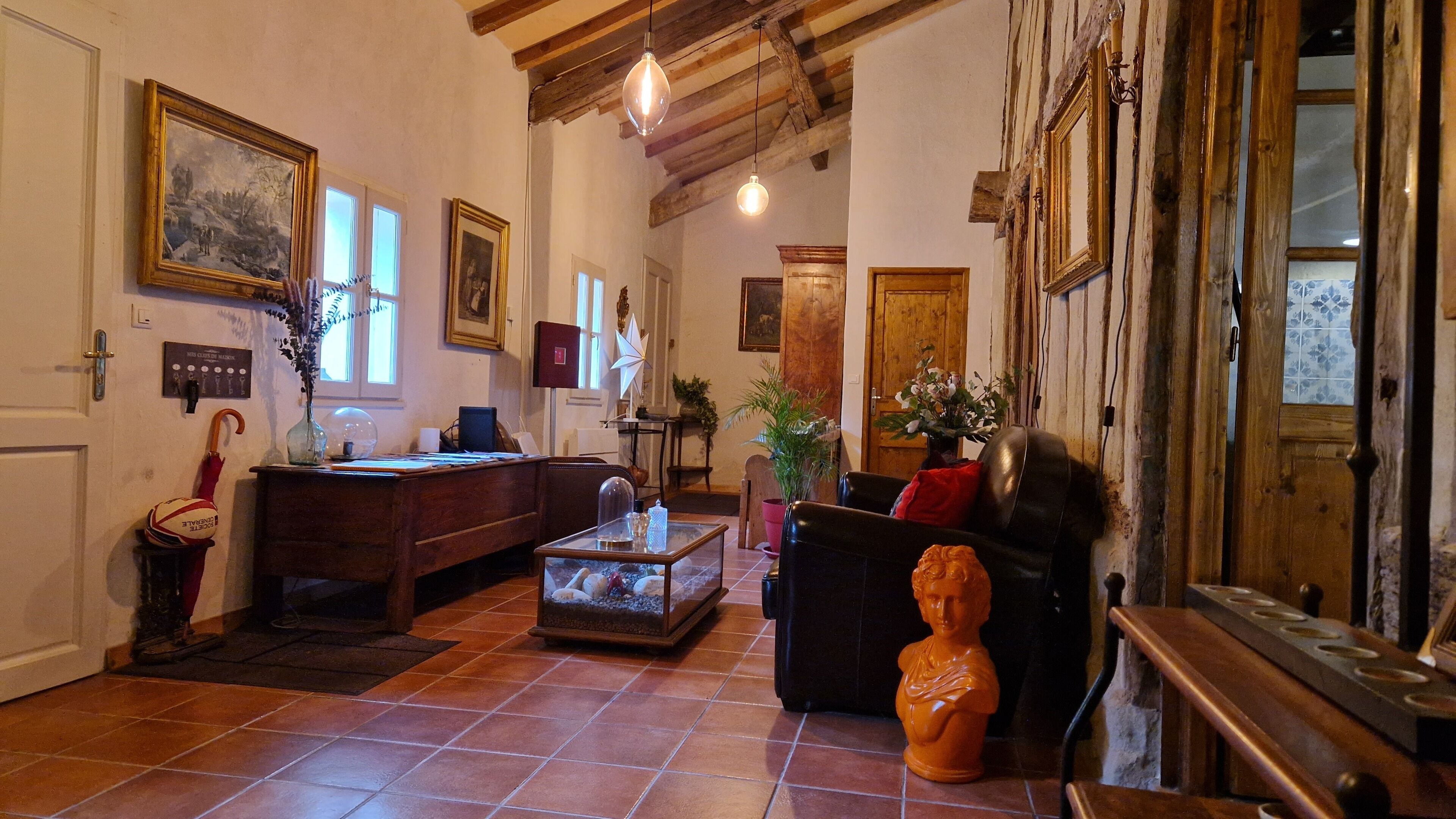 A spacious living room with a wooden desk, a sofa, and a bust sculpture. There are framed pictures on the walls and a large window providing natural light.