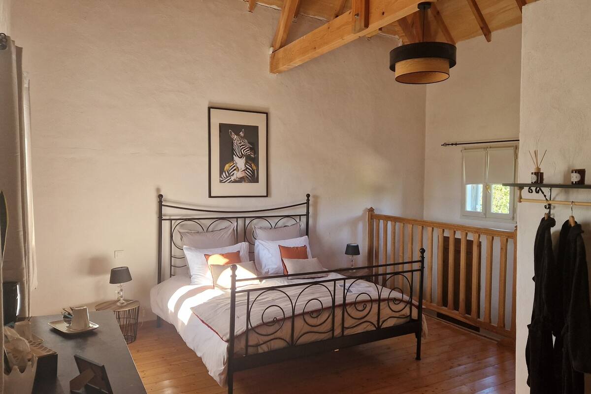 A bedroom with a metal bed frame, a bedside table with a lamp, a framed artwork on the wall, and a wooden ceiling.