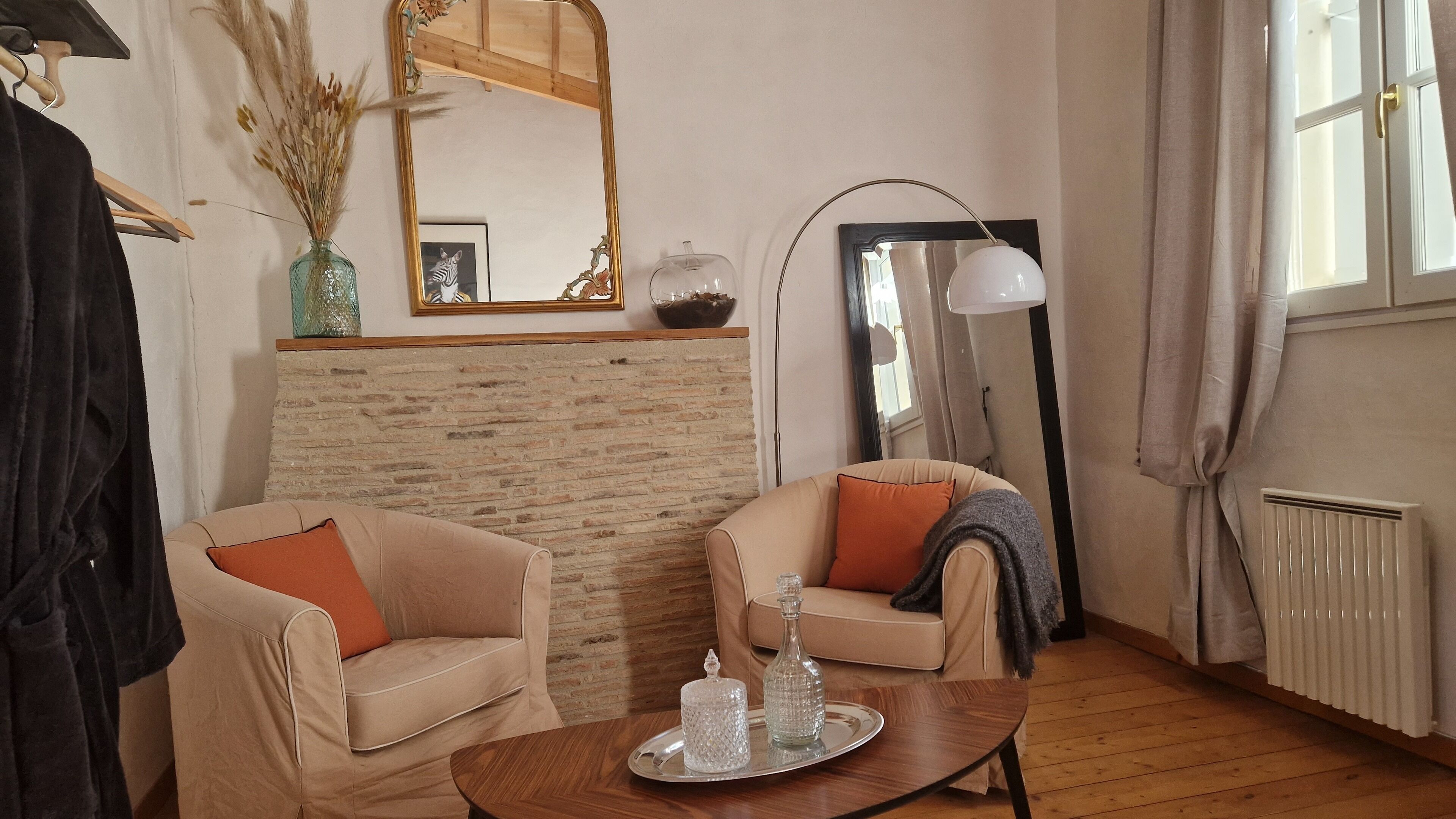 Een gezellige woonkamer met twee beige fauteuils, een houten salontafel met dienblad, een open haard, een grote spiegel en een radiator.