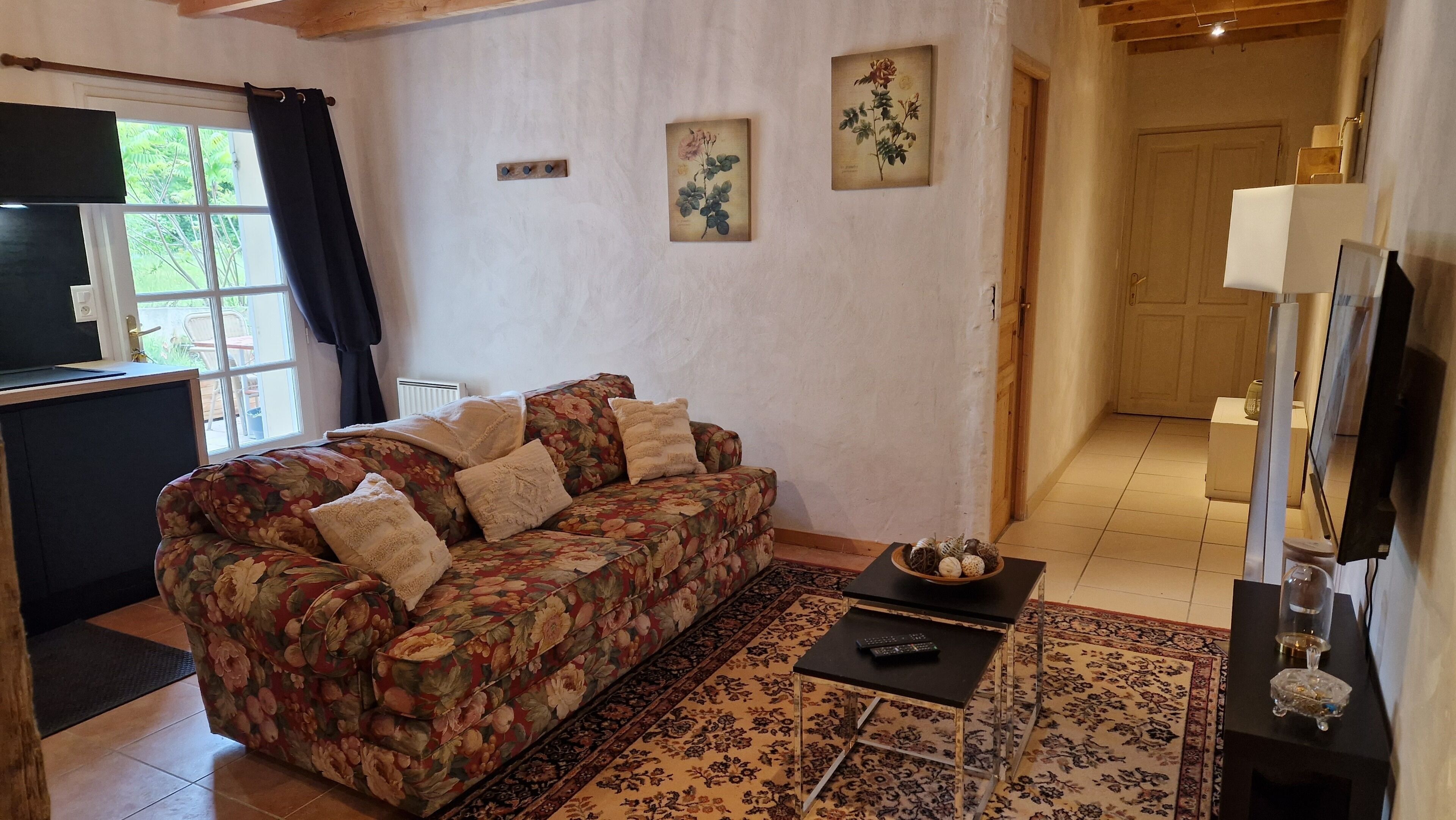 Una sala de estar con un sofá floral, una mesa de centro con objetos decorativos, un televisor y una puerta que da a otra habitación.