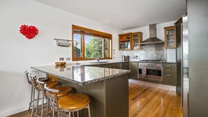 Une cuisine moderne dotée d’un îlot central, de tabourets de bar, d’appareils électroménagers en acier inoxydable et offrant une vue sur la verdure environnante.