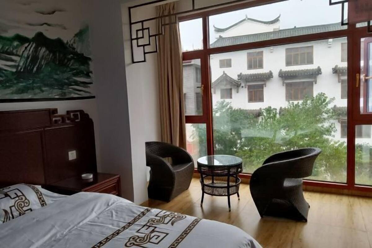 Habitación de hotel con cama, una mesita, dos sillas y un amplio ventanal con vistas a edificios tradicionales y zonas verdes.